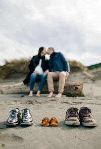 Close-up of shoes with pregnant couple kissing while sitting on land against sky