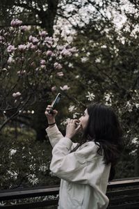 Young woman photographing with mobile phone while standing by tree