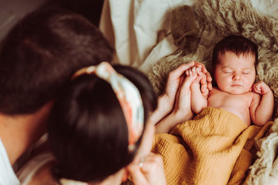 High angle of unrecognizable young mother and father with dark hair holding hand of adorable newborn baby lying and sleeping on soft bed in daylight