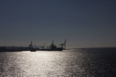 Silhouette cranes by sea against clear sky