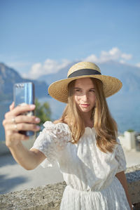 Young woman photographing with mobile phone against sky