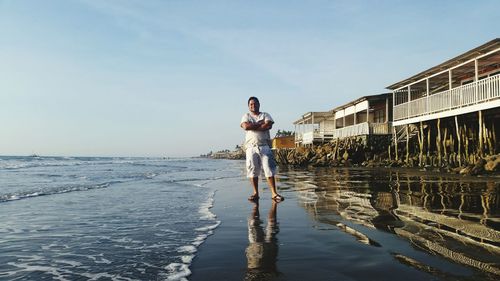 Mid adult man with arms crossed standing at beach against sky during sunset