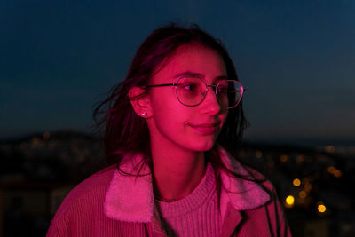 Attentive teen female with long brown hair in eyeglasses admiring dark night in pink illumination