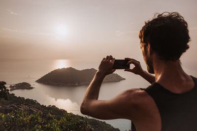 Rear view of woman photographing lake against sky during sunset