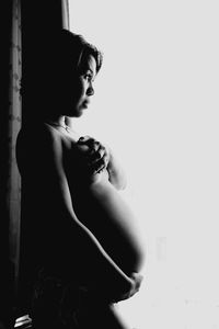 Side view of naked pregnant woman with hand on breast standing by window