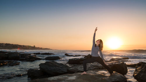 Woman exercising yoga on rock by sea against sky during sunrise