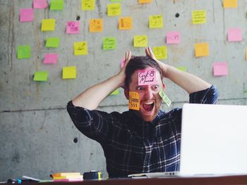 Shocked man with adhesive notes on face sitting by laptop in office