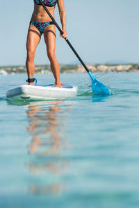 Crop anonymous female surfer standing on surfboard and rowing with paddle while practicing on surface of sea in summer