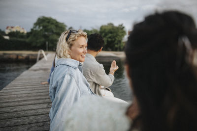 Smiling woman looking away while sitting with friend on pier near lake