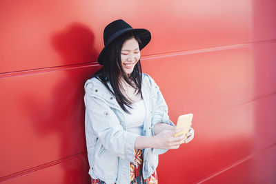 Young woman using mobile phone while standing against red wall