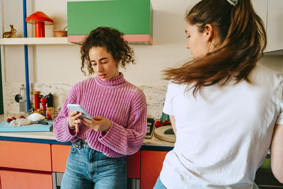 Young woman using smart phone while leaning on kitchen counter by female friend at home