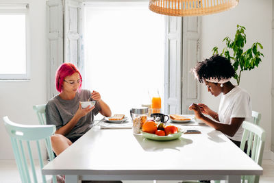 Relaxed young woman with dyed hair sitting near table and eating yummy dish near african american friend in cozy apartment