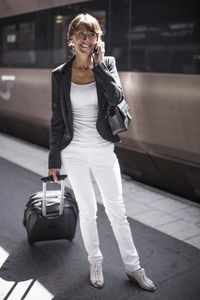 Happy businesswoman with suitcase using mobile phone on railroad station platform