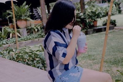 Side view of young woman drinking smoothie while sitting against plants in park