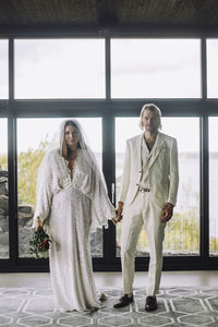 Portrait of mid adult couple holding hands standing against glass door at wedding