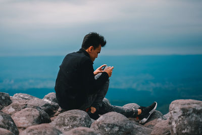 Young man sitting on rocks by sea against sky