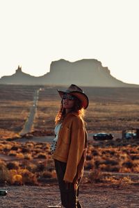 Woman wearing hat standing on land against sky during sunset