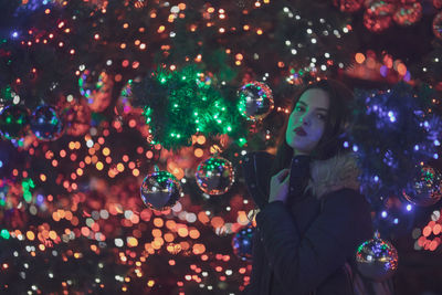 Portrait of young woman standing amidst illuminated christmas tree at night