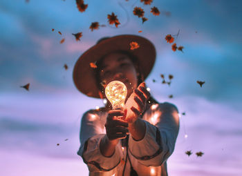 Low angle view of woman holding glowing light bulb amidst falling flowers against sky