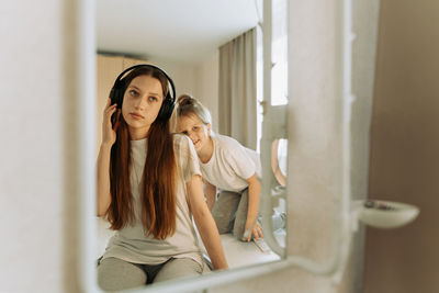 Teenage girl in headphones at mirror, little sister interferes, grimaces.