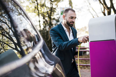 Smiling businessman holding mobile phone at electric vehicle charging station