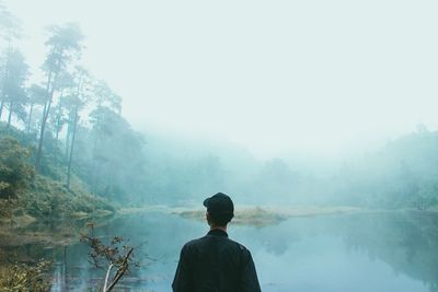 Rear view of man standing by lake against sky during foggy weather
