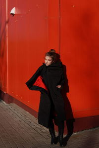 Fashionable teenage girl wearing black warm clothing standing against red wall during sunny day