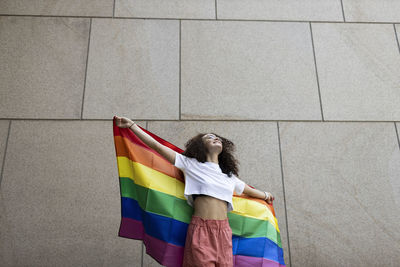 Woman holding rainbow flag while standing in front of wall