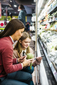 Mother holding artichoke while daughter using mobile phone in supermarket
