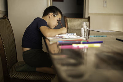 Boy with glasses draws in album with colored pencils, eye fatigue. glass of clean water on the table