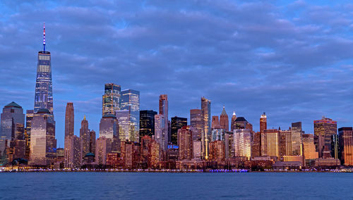 Illuminated buildings in city at waterfront against cloudy sky