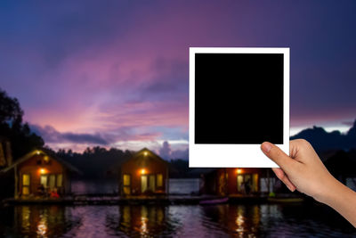 Cropped hand of woman holding blank photograph against sky at dusk