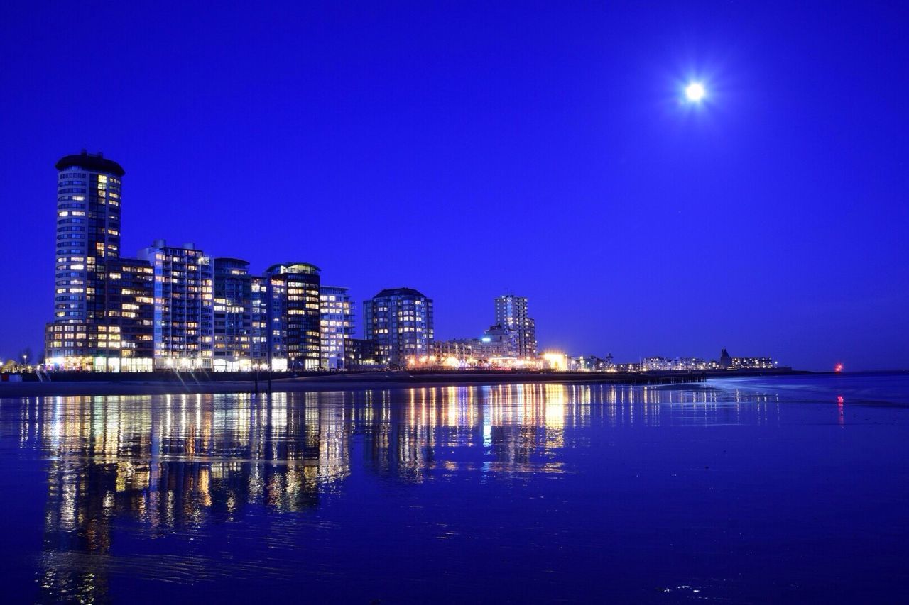 illuminated, night, water, waterfront, reflection, clear sky, blue, architecture, building exterior, built structure, city, copy space, sea, cityscape, moon, sky, river, light, no people, outdoors