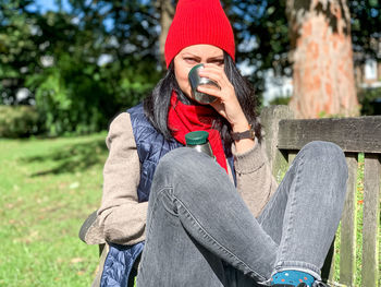 Young millennial woman in casual autumnal clothes siting on wooden bench in park and drinking coffee