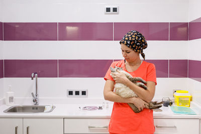 Young female veterinarian in orange uniform holding and hugging cute cat in modern vet clinic