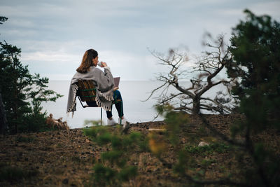 Woman sitting on field by trees against sky