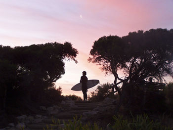 Low angle view of person holding surfboard on steps against sky during sunset