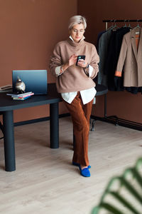 Fashion woman in brown oversize sweater and trousers standing in modern work place with smartphone