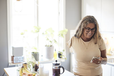 Smiling mature woman with disability using smart phone while standing in kitchen at home