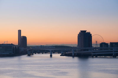 Bridge over river by buildings against sky at sunset