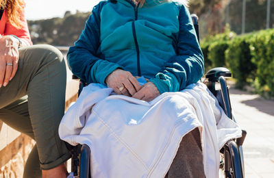 Crop unrecognizable female caregiver sitting with senior woman in wheelchair on sunny day