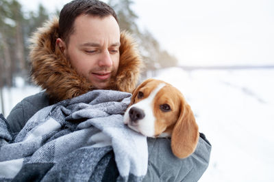 Mid adult man carrying dog in blanket while standing on snow covered field against sky