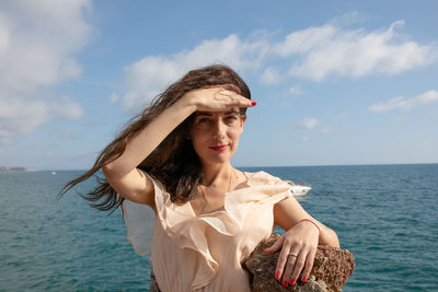 Portrait of woman shielding eyes while standing by rock at beach against sky