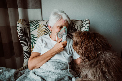 Senior woman wearing oxygen mask sitting with dog on chair at home