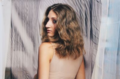 Portrait of beautiful woman looking over shoulder against curtain