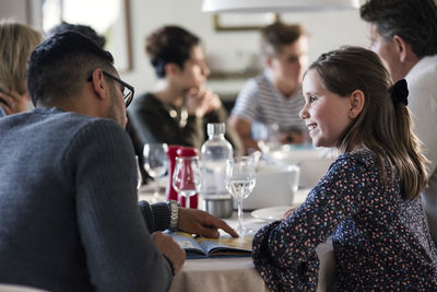 Smiling girl looking at man reading magazine while sitting with friends in dinner party