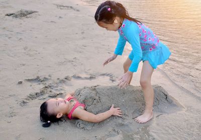 High angle view of girl burying sister with sand at shore of beach