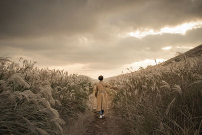 Rear view of mature woman walking on field amidst plants against cloudy sky during sunset