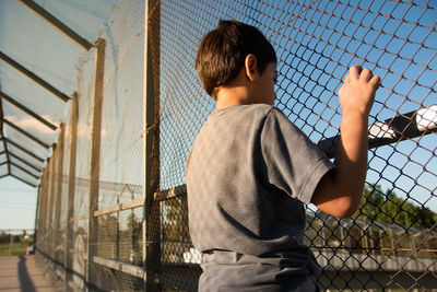 Rear view of boy looking through chainlink fence