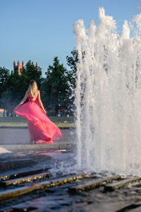 Rear view young woman in pink evening gown standing by fountain in park